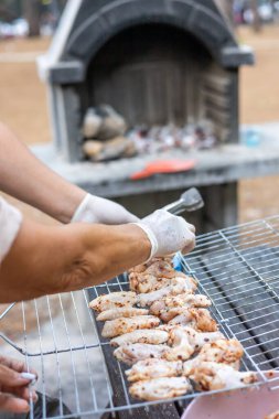 Açık tenli ve eldivenli biri barbeküde tavuk ızgara yapar. Sahne arka planda taş bir ızgarayla dışarıda çekiliyor.