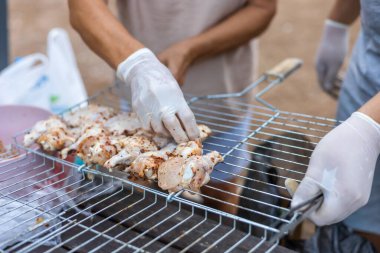 Eller ızgaraya salamura edilmiş tavuk parçaları yerleştiriyor. Sahne, barbekü için hazırlanan yiyeceklerin yakın çekimini gösteriyor. Eldivenler hijyen için giyilir..