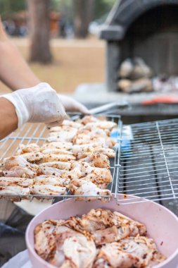 Barbeküde ızgara tavuk. Beyaz eldiven giyen biri ızgarada marine edilmiş tavuk parçaları tutuyor. Parkta açık hava yemek sahnesi.