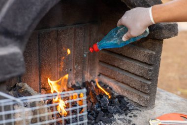 Eldivenli bir el, mangal ızgarasında yanan kömürün üzerine mavi sıvı döküyor. Alevler kömürden yükseliyor, bu da yemek pişirmeye hazırlandığını gösteriyor..