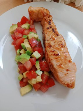 Sağlıklı kahvaltı, öğle yemeği, hafif akşam yemeği. Domatesli ızgara somon ve beyaz tabakta avacado salatası..