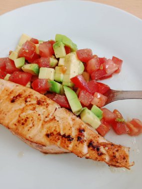 Sağlıklı kahvaltı, öğle yemeği, hafif akşam yemeği. Domatesli ızgara somon ve beyaz tabakta avacado salatası..