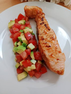 Sağlıklı kahvaltı, öğle yemeği, hafif akşam yemeği. Domatesli ızgara somon ve beyaz tabakta avacado salatası..