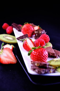 Meyve tabağı. Dikdörtgen bir tabakta çilek, ahududu, kivi ve çikolata parçaları.