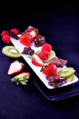 Meyve tabağı. Dikdörtgen bir tabakta çilek, ahududu, kivi ve çikolata parçaları.