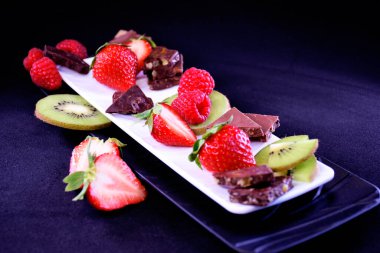 Meyve tabağı. Dikdörtgen bir tabakta çilek, ahududu, kivi ve çikolata parçaları.