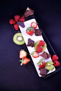 Meyve tabağı. Dikdörtgen bir tabakta çilek, ahududu, kivi ve çikolata parçaları.