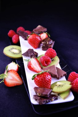Meyve tabağı. Dikdörtgen bir tabakta çilek, ahududu, kivi ve çikolata parçaları.