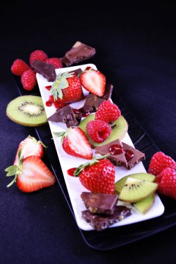 Meyve tabağı. Dikdörtgen bir tabakta çilek, ahududu, kivi ve çikolata parçaları.