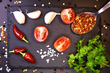 Siyah bir mat yüzeyinde yemek pişirmek için gerekli malzemeler. Ön planda parlak kırmızı biberler, olgun domates parçaları ve tahta üzerine yerleştirilmiş sarımsak karanfilleri uzanıyor. Çevrelerinde, sert deniz tuzu, kuru kırmızı biber taneleri, siyah beyaz biber taneleri vardır..