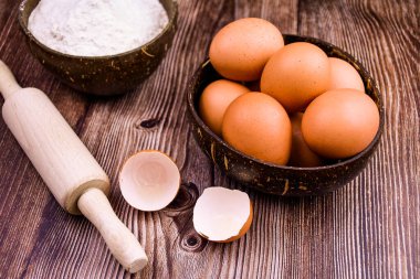 Tahta bir masanın arka planında, ahşap bir kasede tavuk yumurtası. Ön planda iki kırık yumurta kabuğu, bir kasede un ve ahşap bir oklava. Mutfakta sade bir atmosfer yaratmak için kompozisyon, pişirme...