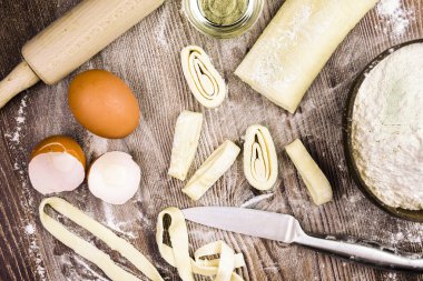 Ev yapımı makarna yapmak için malzemeler: yumurta, rulo hamur, bir kasede un, rulo şeklinde kesilmiş hamur parçaları, bir bıçak ve merdane. Bu kompozisyon aşçılık yaratıcılığı sürecinde bir his yaratıyor..