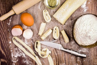 Ev yapımı makarna yapmak için malzemeler: yumurta, rulo hamur, bir kasede un, rulo şeklinde kesilmiş hamur parçaları, bir bıçak ve merdane. Bu kompozisyon aşçılık yaratıcılığı sürecinde bir his yaratıyor..