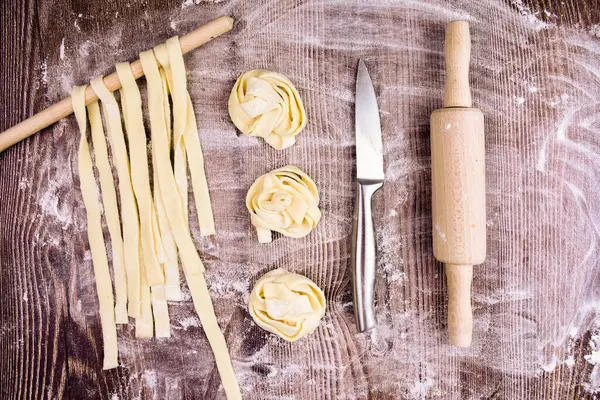 Ev yapımı makarna yapmak için malzemeler: yumurta, rulo hamur, bir kasede un, rulo şeklinde kesilmiş hamur parçaları, bir bıçak ve merdane. Bu kompozisyon aşçılık yaratıcılığı sürecinde bir his yaratıyor..