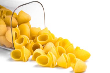 Kabuk şeklindeki makarna kısmen şeffaf cam bir kavanozda kısmen ahşap kapaklı, kısmen beyaz arka plana saçılmış. Hafif arka plan makarnanın dokusunu ve rengini vurguluyor, minimalist ve estetik bir görünüm yaratıyor.. 