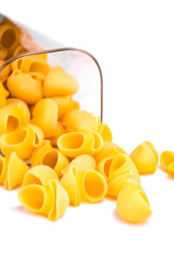 Kabuk şeklindeki makarna kısmen şeffaf cam bir kavanozda kısmen ahşap kapaklı, kısmen beyaz arka plana saçılmış. Hafif arka plan makarnanın dokusunu ve rengini vurguluyor, minimalist ve estetik bir görünüm yaratıyor.. 