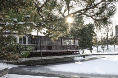 Donmuş bir göletin kıyısında ahşap bir evi olan kış manzarası. Güneş ışınları karla kaplı kalın çam dallarını delip geçer..