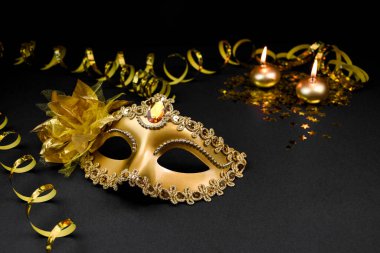 Parıltı ve çiçekle süslenmiş altın bir karnaval maskesi, yüzen bir mum ve altın boncuklu bir çelenk içeren siyah bir arkaplanda kompozisyon..