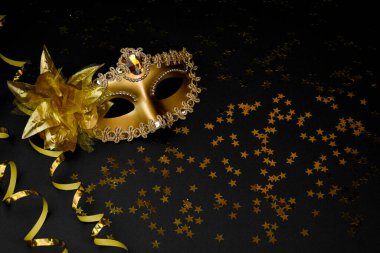 Parıltı ve çiçekle süslenmiş altın bir karnaval maskesi, yüzen bir mum ve altın boncuklu bir çelenk içeren siyah bir arkaplanda kompozisyon..