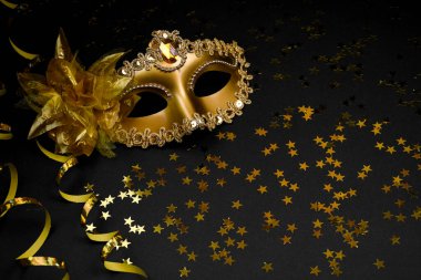 Parıltı ve çiçekle süslenmiş altın bir karnaval maskesi, yüzen bir mum ve altın boncuklu bir çelenk içeren siyah bir arkaplanda kompozisyon..