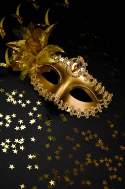 Parıltı ve çiçekle süslenmiş altın bir karnaval maskesi, yüzen bir mum ve altın boncuklu bir çelenk içeren siyah bir arkaplanda kompozisyon..