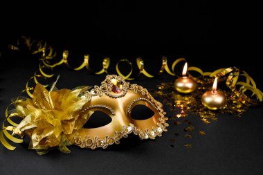 Parıltı ve çiçekle süslenmiş altın bir karnaval maskesi, yüzen bir mum ve altın boncuklu bir çelenk içeren siyah bir arkaplanda kompozisyon..