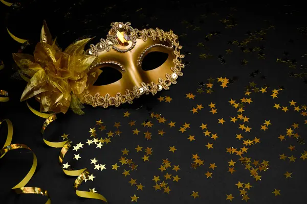 Parıltı ve çiçekle süslenmiş altın bir karnaval maskesi, yüzen bir mum ve altın boncuklu bir çelenk içeren siyah bir arkaplanda kompozisyon..