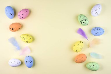 Colorful motley Easter eggs located along the edges of the image on a pastel yellow background with soft decorative feathers. clipart