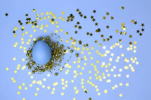Altın boncuklarla süslenmiş mavi bir Paskalya yumurtası ve yumuşak mavi bir arka planda yıldız konfetisi.