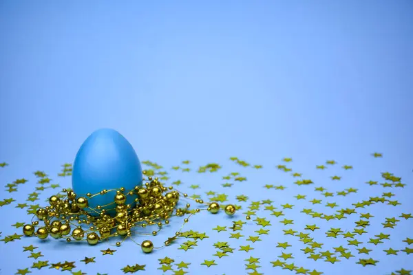 Altın boncuklarla süslenmiş mavi bir Paskalya yumurtası ve yumuşak mavi bir arka planda yıldız konfetisi.