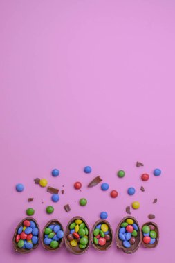 Renkli çikolata kaplı kırık çikolata yumurtaları pastel pembe arka planda düzenlenir. Küçük şekerler ve çikolata parçaları etrafa saçılmış.