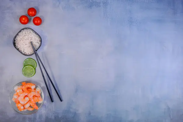 Asya yemeği pişirmek için malzemelerle bezenmiş düz bir kompozisyon. Arka planda cam kasedeki karidesler, siyah seramik kasedeki beyaz pirinç, taze limon dilimleri, vişneli domatesler ve siyah çubuklar var..