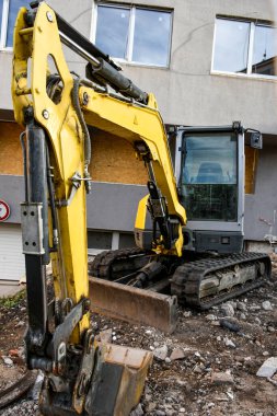 Modern bir binanın yakınındaki bir şantiyede kompakt kazıcı. Kentsel gelişim ve yenileme çalışmaları için kullanılan ağır makineler. Arka planda bina cephesi, ön planda moloz ve kir.