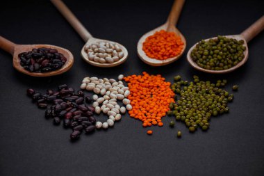 Tahta kaşıklarda çeşitli baklagiller var ve siyah arka plana saçılmışlar. Sağlıklı bitkisel besin maddeleri. Yüksek proteinli vegan ürünleri.