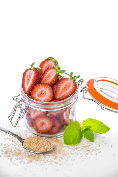 Dilimlenmiş taze çilekli cam kavanoz, esmer şeker ve beyaz arka planda nane yaprakları. Yaz tatlısı malzemeleri. Temiz yeme konsepti. Dikey bileşim.