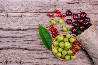 Taze yaz meyveleri - kiraz, bektaşi üzümü ve kırmızı üzümler - ahşap arka planda yeşil yapraklar ve keten poşetlerle düzenlenmiş. Mevsimlik meyveler