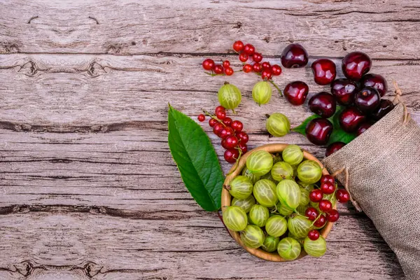 Taze yaz meyveleri - kiraz, bektaşi üzümü ve kırmızı üzümler - ahşap arka planda yeşil yapraklar ve keten poşetlerle düzenlenmiş. Mevsimlik meyveler