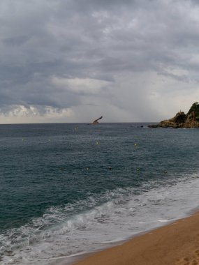 Deniz dalgaları kumlu sahilde, kayalara vuruyor. Denizde bir yelkenli. Mavi denizin üzerinde bir güvercin gökyüzü. Yüksek kalite fotoğraf