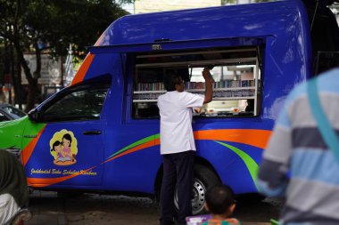 Malang şehrinin ortasında bir okur yazarlık alanında bir araba (mobil kütüphane) kitap rafını düzenleyen bir adam