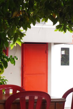 Beyaz binadaki kırmızı demir kapı. Yapraklar çerçeveyi ve kırmızı plastik sandalyeleri oluşturuyor.