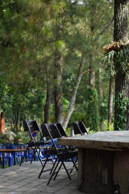 Gölgeli bir günde arka planda ağaçlar olan bir kafede düzgün düzenlenmiş siyah sandalyeli uzun tahta bir masa.