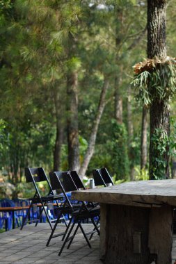 Gölgeli bir günde arka planda ağaçlar olan bir kafede düzgün düzenlenmiş siyah sandalyeli uzun tahta bir masa.