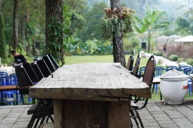 Gölgeli bir günde arka planda ağaçlar olan bir kafede düzgün düzenlenmiş siyah sandalyeli uzun tahta bir masa.