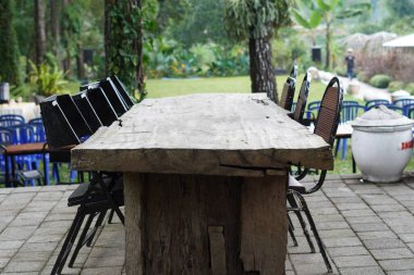 Gölgeli bir günde arka planda ağaçlar olan bir kafede düzgün düzenlenmiş siyah sandalyeli uzun tahta bir masa.