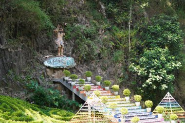 Gün boyunca Taman Kemesraan, Pujon, Batu, Malang 'da ağaçlı bir tepede bir tanrı heykeline giden renkli adımlar.