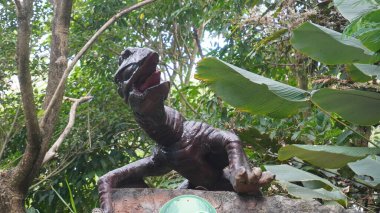 A dinosaur statue that is opening its mouth and looking to the side against a background of leaves and trees clipart