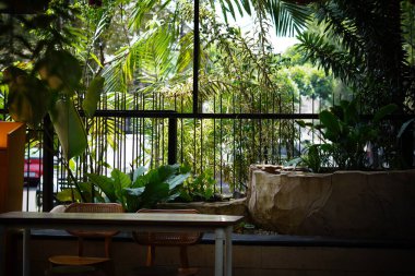 Kafenin arka planında yeşil bir bitki olan tahta sandalyeler ve masalar arasında oturan taş koltuklar.