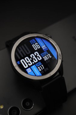 A black smartwatch that is on and displays date and time and health information paired with a smartphone on a black background clipart