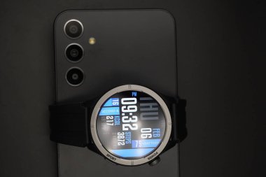 A black smartwatch that is on and displays date and time and health information paired with a smartphone on a black background clipart