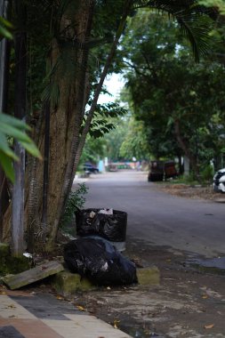 Çöp dolu büyük siyah bir plastik torba ağaçlı sessiz bir otobanın yanında yer almaktadır.